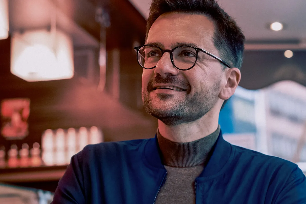 Thomas Kutschaty lächelt mit Brille und blauer Jacke über einem braunen Rollkragenpullover.