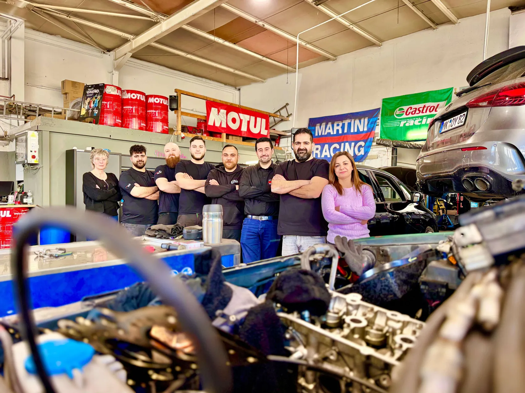 Sieben Personen stehen in einer Autowerkstatt vor Regalen mit Autozubehör und einem demontierten Motor. Ahmet Tümce, der Inhaber, steht mittig mit verschränkten Armen und seinem Team.