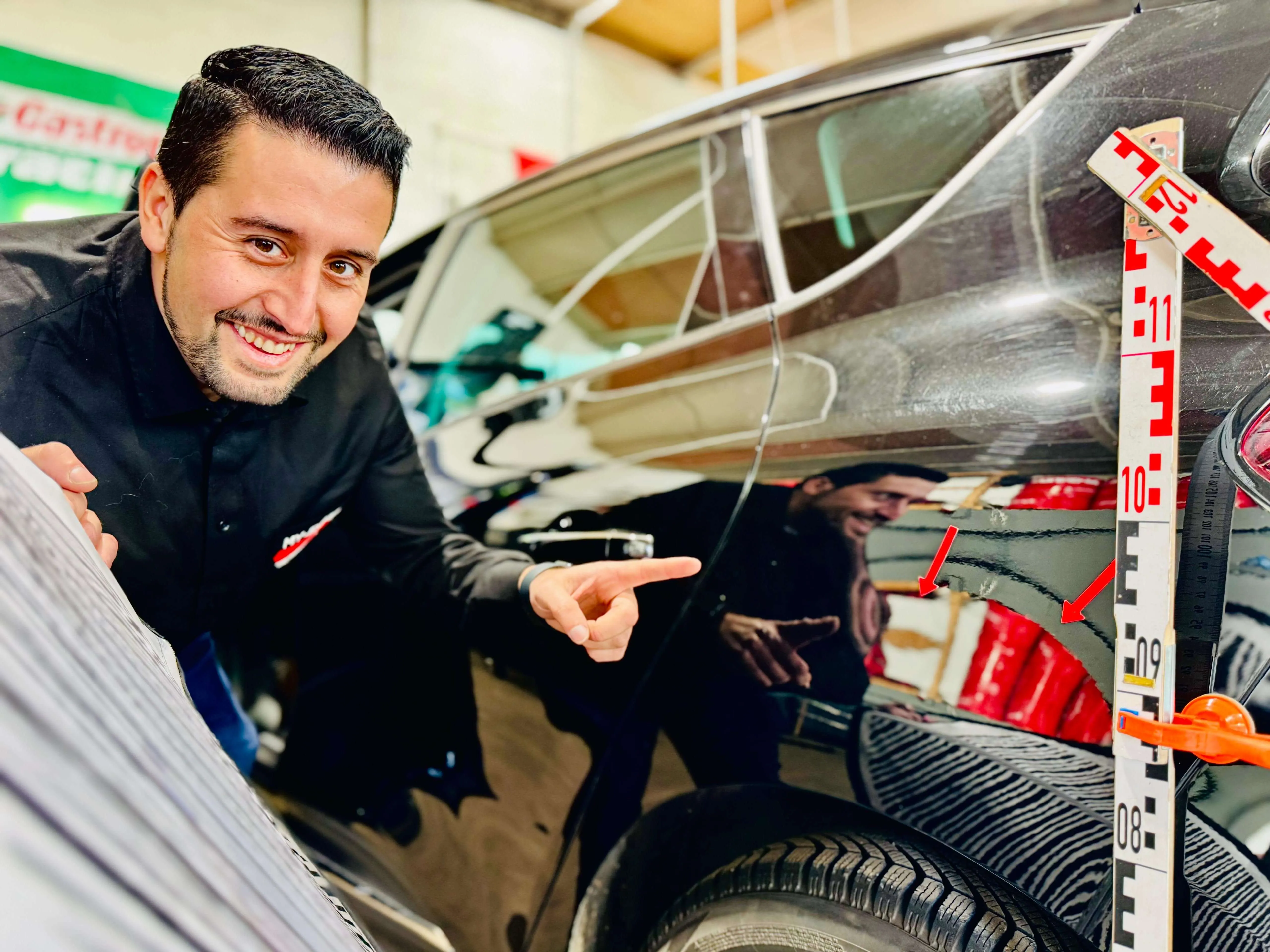 Ein lächelnder Mann in Arbeitskleidung zeigt auf einen Schaden am Kotflügel eines grauen Autos, während er eine Messlatte hält.