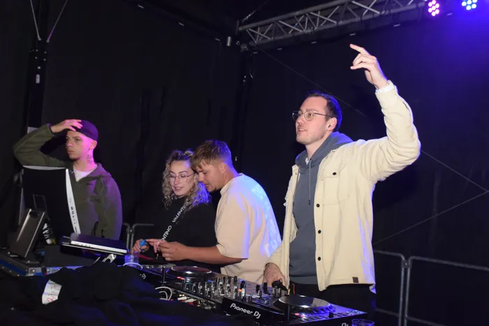 DJ mit erhobener Hand vor einem DJ-Pult mit mehreren Personen im Hintergrund.