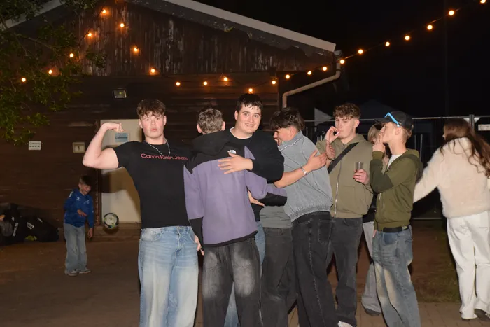 Eine Gruppe junger Leute umarmt und posiert lachend vor einem Holzgebäude, das mit Lichterketten dekoriert ist. Im Hintergrund tanzt ein Kind.