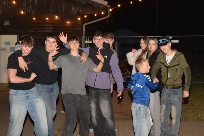 Eine Gruppe junger Menschen posiert lächelnd und gestikulierend vor einer dunklen Kulisse mit Lichterketten. Einige haben ihre Arme um die Schultern der anderen gelegt.