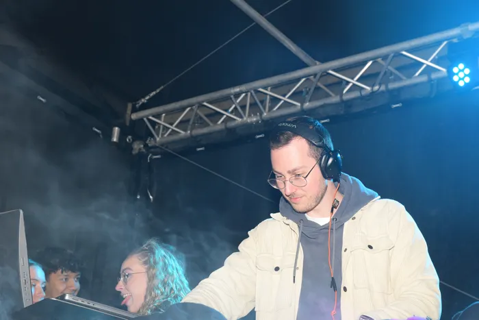 Ein DJ mit Kopfhörern steht hinter den Plattenspielern und legt Musik auf. Im Hintergrund ist eine tanzende Person und Bühnentechnik zu sehen.