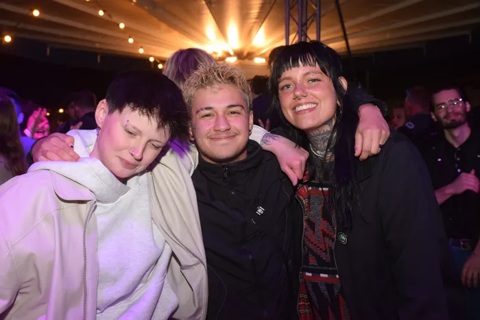 Drei junge Erwachsene umarmen sich lachend vor einer Menschenmenge auf einer Party. Die Person rechts hat mehrere Piercings und Tattoos im Gesicht und am Hals.