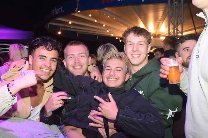 Vier junge Männer lächeln in die Kamera, eng aneinander geschmiegt, während im Hintergrund eine Menschenmenge auf einer Party tanzt und sich unterhält. Einer der Männer hält ein Bierglas in der Hand.