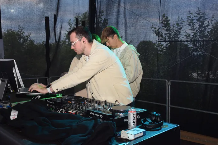 Zwei DJs spielen vor Publikum auf. Der DJ links trägt eine Brille und eine helle Jacke, während der DJ rechts eine Kappe trägt.
