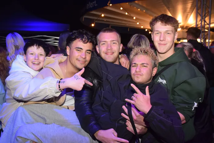 Fünf junge Erwachsene posieren eng zusammen und lächeln in die Kamera. Im Hintergrund ist eine Menschenmenge und eine beleuchtete Zeltkonstruktion zu sehen.