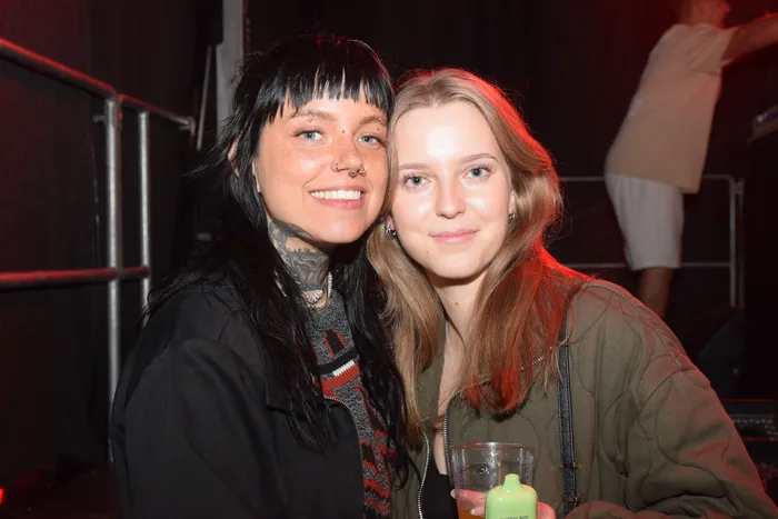 Zwei lächelnde junge Frauen posieren eng nebeneinander. Die linke Person hat dunkle Haare, mehrere Piercings und Tattoos, während die rechte Person hellere Haare hat und ein Getränk in der Hand hält. Im Hintergrund sind weitere Menschen und eine metallene Barriere zu sehen.