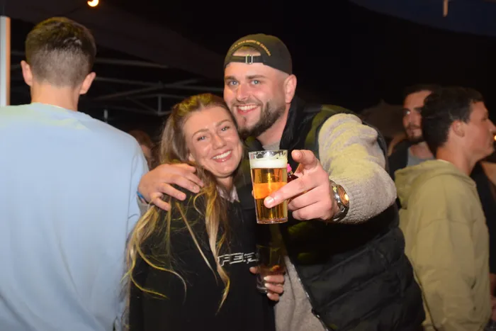 Ein lächelnder Mann mit Kappe hält ein Bierglas hoch, während eine Frau ihn von der Seite umarmt. Im Hintergrund sind weitere feiernde Menschen zu sehen.