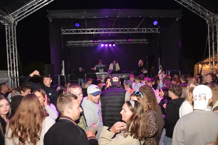 Eine große Menschenmenge tanzt vor einer Bühne mit einem DJ, der Musik auflegt. Die Party findet im Freien statt und ist mit violettem Licht beleuchtet.