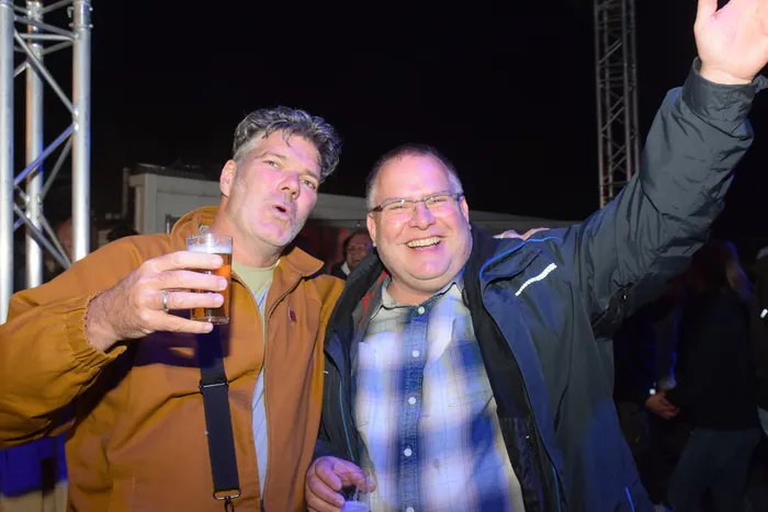 Zwei lächelnde Männer stehen eng zusammen, einer hält ein Bierglas, der andere hebt die Hand. Im Hintergrund ist eine beleuchtete Bühne und tanzende Menschen zu sehen.