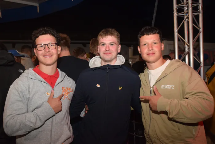 Drei junge Männer stehen eng zusammen und schauen in die Kamera. Sie tragen Freizeitkleidung und lächeln. Im Hintergrund sind weitere Menschen auf einer Party zu sehen.