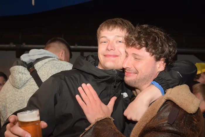 Zwei junge Männer umarmen sich eng, während ein Mann im Hintergrund einen Bierkrug hält. Die Szene zeigt ausgelassene Stimmung auf einer Party.