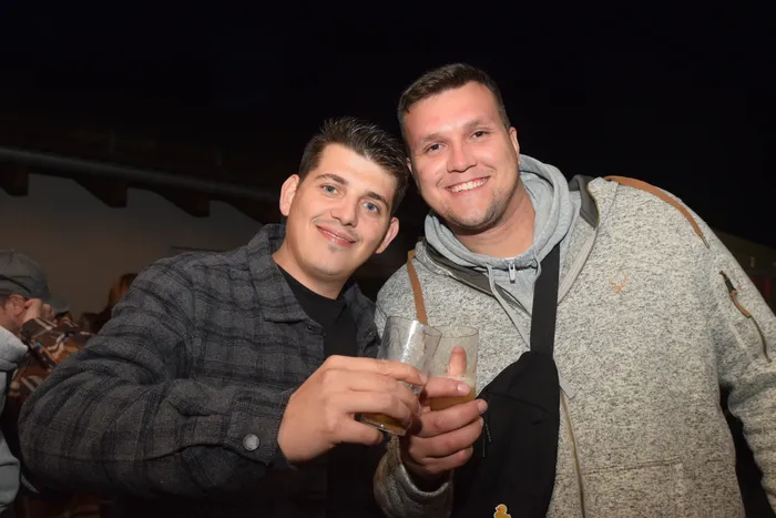 Zwei junge Männer lächeln in die Kamera und halten jeweils ein Glas mit Bier in der Hand. Im Hintergrund sind weitere Menschen auf einer Party zu sehen.