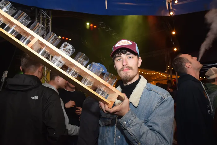 Ein junger Mann mit roter Kappe und Jeansjacke balanciert ein Tablett mit vollen Biergläsern auf der Schulter, umgeben von feiernden Menschen. Im Hintergrund ist eine Bühne mit Lichtinstallationen zu sehen.