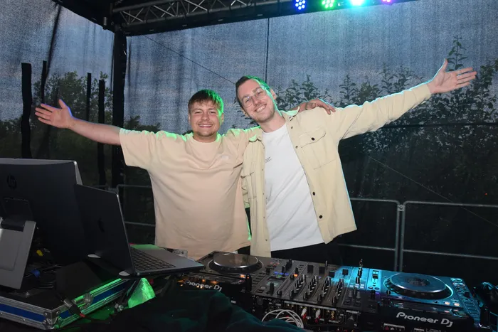 Zwei junge Männer stehen lächelnd vor einem DJ-Pult mit weit ausgebreiteten Armen. Im Hintergrund ist eine Bühne und eine tanzende Menschenmenge zu erkennen.
