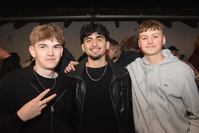 Drei junge Männer posieren eng nebeneinander für ein Foto. Der Mann in der Mitte lächelt in die Kamera, während die beiden anderen leicht lächeln. Im Hintergrund sind weitere Menschen auf einer Party zu sehen.
