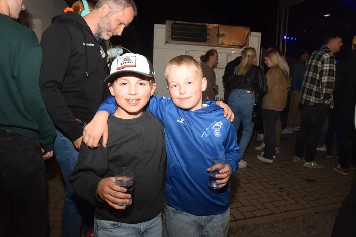 Zwei lächelnde Jungen mit Getränken im Glas stehen eng umschlungen vor einer Menschenmenge auf einer nächtlichen Party. Im Hintergrund ist ein Transporter und weitere feiernde Personen zu sehen.
