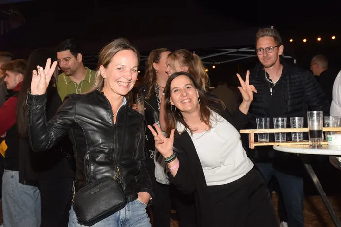 Zwei lächelnde Frauen zeigen beim Feiern auf einer Party das Victory-Zeichen. Im Hintergrund tanzen und stehen weitere Menschen, Getränke werden serviert.