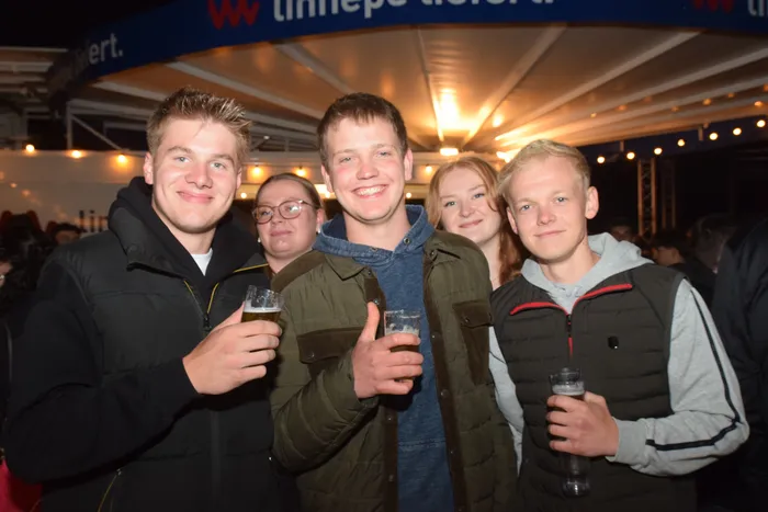Eine Gruppe junger Erwachsener lächelt in die Kamera, einige halten Biergläser in der Hand. Im Hintergrund ist ein beleuchtetes Zelt mit Menschen zu sehen.