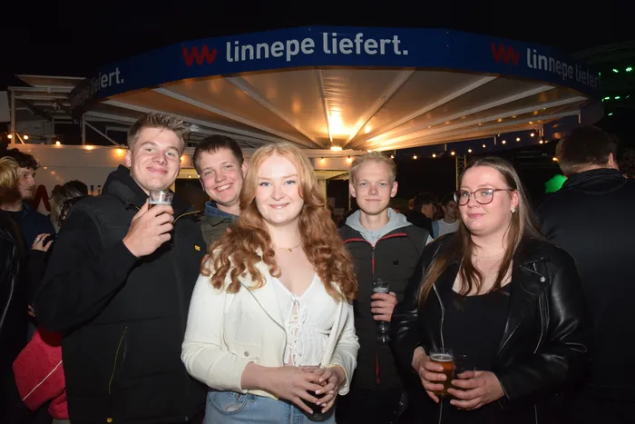 Fünf junge Erwachsene stehen lächelnd vor der Kamera und halten Biergläser in der Hand. Im Hintergrund ist ein Zelt mit der Aufschrift „linnepe liefert.“ zu sehen.