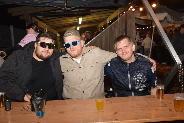 Drei junge Männer lächeln in die Kamera, stehen eng nebeneinander an einer Theke und halten Biergläser in der Hand. Im Hintergrund ist eine belebte Party mit weiteren Menschen zu sehen.