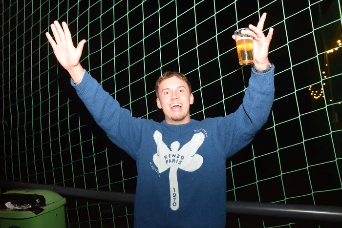 Ein junger Mann mit hoch erhobenen Armen und einem Bierglas in der Hand steht vor einem grünen Gitter. Er lächelt und scheint die ausgelassene Stimmung auf der Party zu genießen.