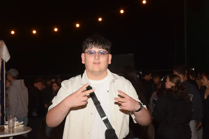 Junge Person mit dunklen Haaren, Brille und offener Jacke zeigt mit beiden Zeigefingern nach links und rechts in die Menge auf einer Party. Im Hintergrund sind weitere Menschen und Lichterketten zu sehen.