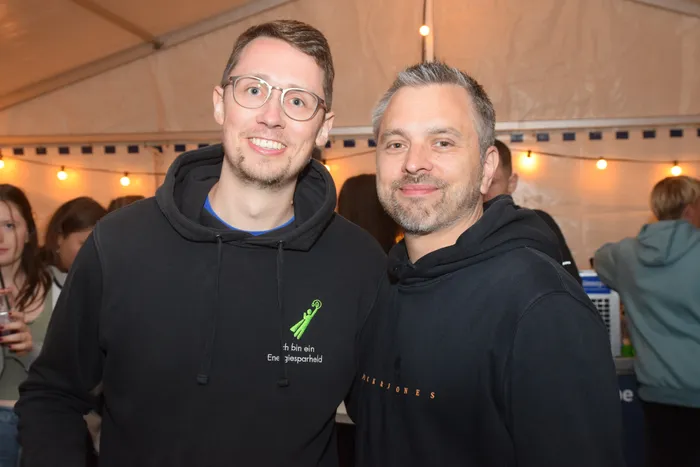 Zwei lächelnde Männer posieren eng nebeneinander für das Foto. Beide tragen dunkle Kapuzenpullover, der linke zeigt einen grünen Aufdruck mit der Aufschrift „Ich bin ein Energiesparheld“. Im Hintergrund sind weitere Menschen und festliche Beleuchtung zu sehen.