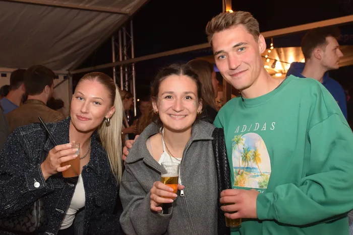 Drei junge Erwachsene stehen lächelnd eng zusammen und halten jeweils ein Glas mit Getränk in der Hand. Im Hintergrund sind weitere Menschen auf einer Party zu sehen.