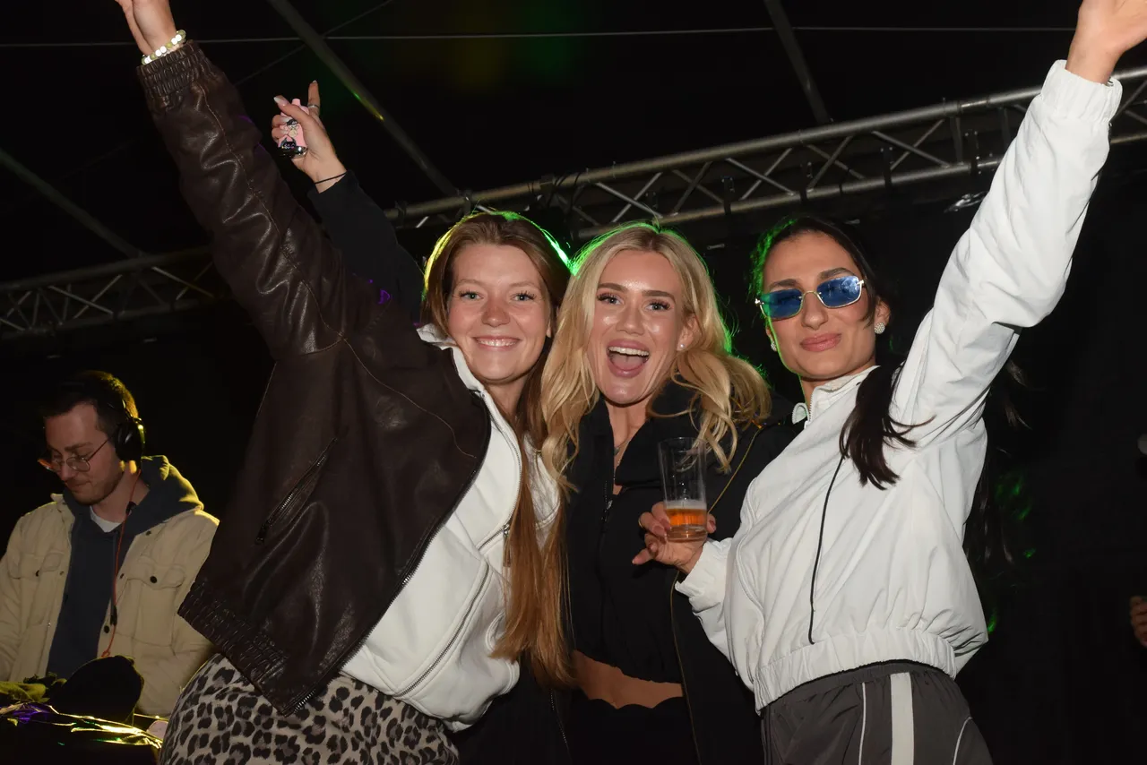 Drei junge Frauen mit hoch erhobenen Armen tanzen vor einer Bühne. Im Hintergrund ist ein DJ zu sehen.