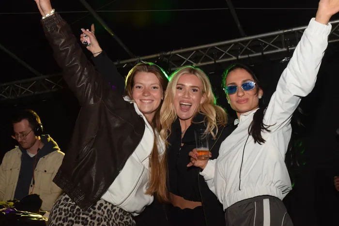 Drei junge Frauen mit hoch erhobenen Armen tanzen vor einer Bühne. Im Hintergrund ist ein DJ zu sehen.