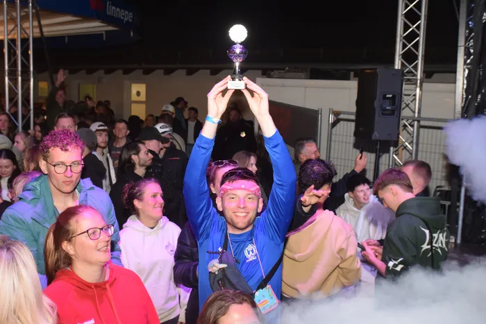 Ein Mann hält eine silberne Trophäe in die Höhe, umgeben von einer jubelnden Menschenmenge auf einer Party im Freien. Nebelmaschinen erzeugen eine stimmungsvolle Atmosphäre.