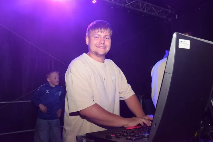 Ein lächelnder junger Mann steht hinter einem DJ-Pult mit Kopfhörern auf und bedient die Geräte. Im Hintergrund ist ein Junge in blauem Trainingsanzug zu sehen. Die Szene ist in violettes Licht getaucht.