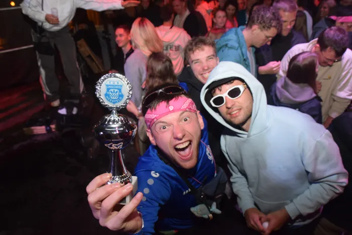 Zwei junge Männer halten einen Pokal in die Kamera, umgeben von feiernden Menschen auf einer Tanzfläche. Der Mann im Vordergrund schreit vor Freude.
