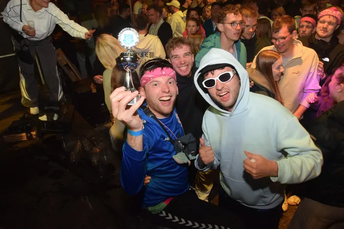 Zwei junge Männer lächeln in die Kamera, einer hält einen Pokal hoch. Im Hintergrund tanzen und feiern viele Menschen auf einer Party.