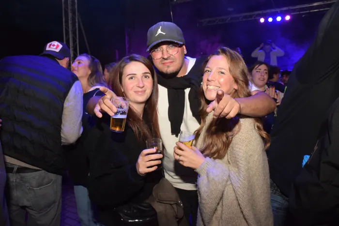 Drei junge Erwachsene lächeln in die Kamera, zwei von ihnen halten Biergläser. Im Hintergrund ist eine tanzende Menschenmenge in einem dunklen Raum mit blauer Beleuchtung zu sehen.