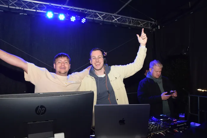 Zwei junge Männer stehen hinter einem DJ-Pult, beide mit erhobenen Armen und lächelnd. Im Hintergrund ist ein dritter Mann und Bühnentechnik zu sehen.