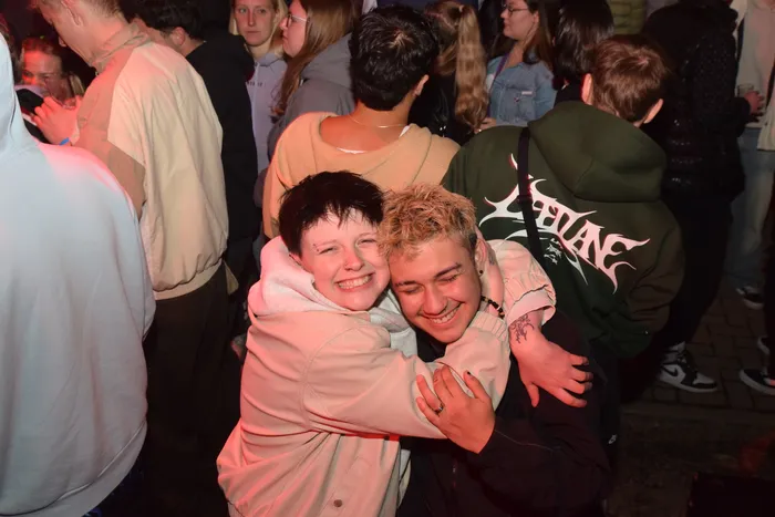 Zwei junge Menschen umarmen sich lachend inmitten einer Menschenmenge auf einer Party. Im Hintergrund sind weitere feiernde Menschen und rotes Licht zu sehen.