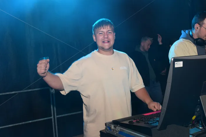 Ein junger Mann mit hochgehobenem Arm steht vor einem DJ-Pult mit Lichteffekten und steuert die Musik bei einer Party. Im Hintergrund sind weitere Personen und eine Tanzfläche zu erkennen.