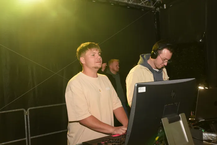 Zwei junge Männer stehen an einem DJ-Pult mit Laptop und Mischpult, während im Hintergrund weitere Personen zu sehen sind. Einer trägt Kopfhörer und konzentriert sich auf seine Arbeit.