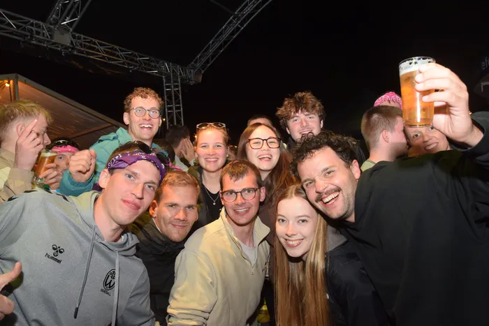 Eine Gruppe junger Menschen posiert eng zusammen und lächelt in die Kamera. Einige halten Bierkrüge hoch, während andere mit Daumen hoch die Stimmung unterstützen. Im Hintergrund ist eine dunkle Umgebung mit Bühnenelementen zu erkennen.