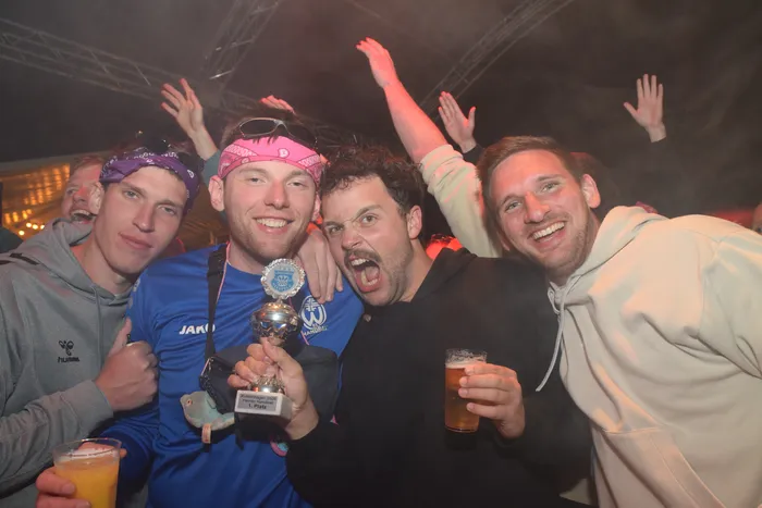Feiernde Menschen umgeben einen Mann mit Medaille und Pokal, alle halten Biergläser in der Hand und lächeln in die Kamera. Im Hintergrund ist eine tanzende Menge und Lichttechnik zu sehen.