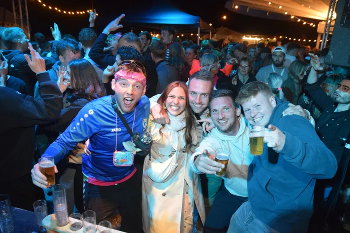 Eine Gruppe junger Erwachsener hält Biergläser in die Kamera, während im Hintergrund eine Menschenmenge auf einer Tanzfläche feiert. Die Atmosphäre ist ausgelassen und festlich beleuchtet.