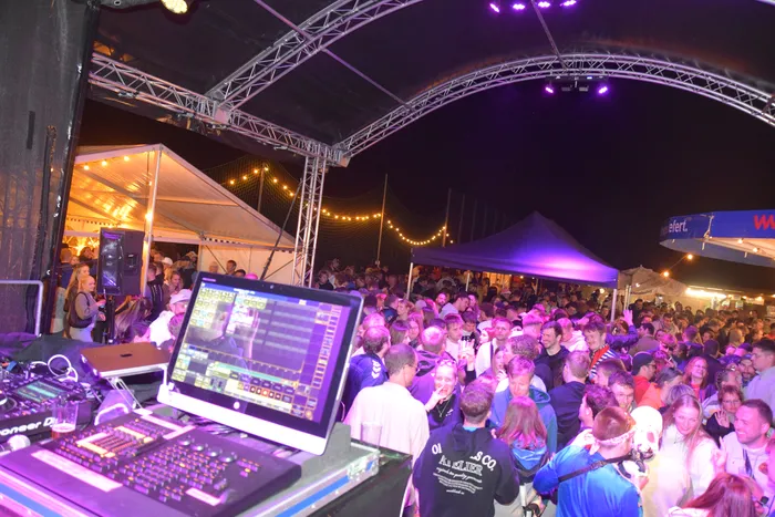 Eine große Menschenmenge tanzt vor einer Bühne bei einem Open-Air-Event in Kuhlenhagen. Im Vordergrund ist ein DJ-Pult mit einem Monitor zu sehen, auf dem Visualisierungen laufen.