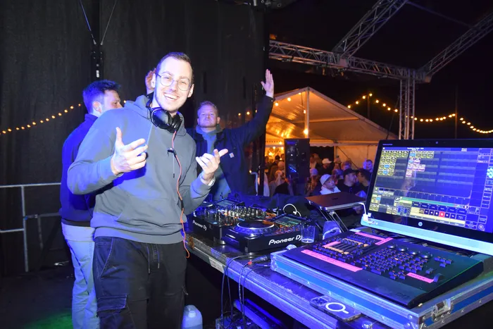 Ein DJ mit Kopfhörern steht hinter einem Mischpult vor einer Menschenmenge auf einer Tanzfläche unter einem Zelt. Er lächelt und hat die Hände erhoben.