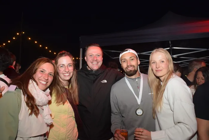 Fünf lächelnde Menschen stehen eng zusammen vor einer dunklen Kulisse mit Partyzelten und Lichterketten. Ein Mann trägt eine Medaille um den Hals und hält ein Bierglas. Im Hintergrund sind weitere feiernde Menschen zu erkennen.