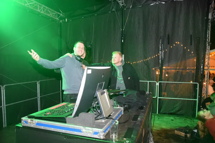 Ein DJ mit Kopfhörern steht hinter einem DJ-Pult mit Laptop und mischt Musik. Im Hintergrund ist ein weiterer Mann zu sehen und eine tanzende Menschenmenge.