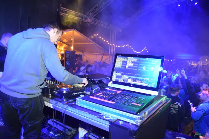 Ein DJ steht hinter einem Mischpult vor einer großen Menschenmenge auf einer Tanzfläche bei einem Open-Air-Event. Auf dem Bildschirm des Mischpults sind Visualisierungen zu sehen. Im Hintergrund sind Lichterketten und ein Zelt zu erkennen.