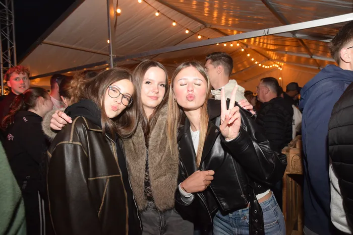 Drei junge Frauen posieren eng umschlungen und lächelnd vor einer Menschenmenge in einem beleuchteten Zelt. Eine von ihnen macht das Victory-Zeichen.